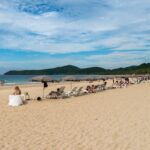 Is Nacpan Beach swimmable? Calm blue water beside a sandy beach with umbrellas and lounge chairs.