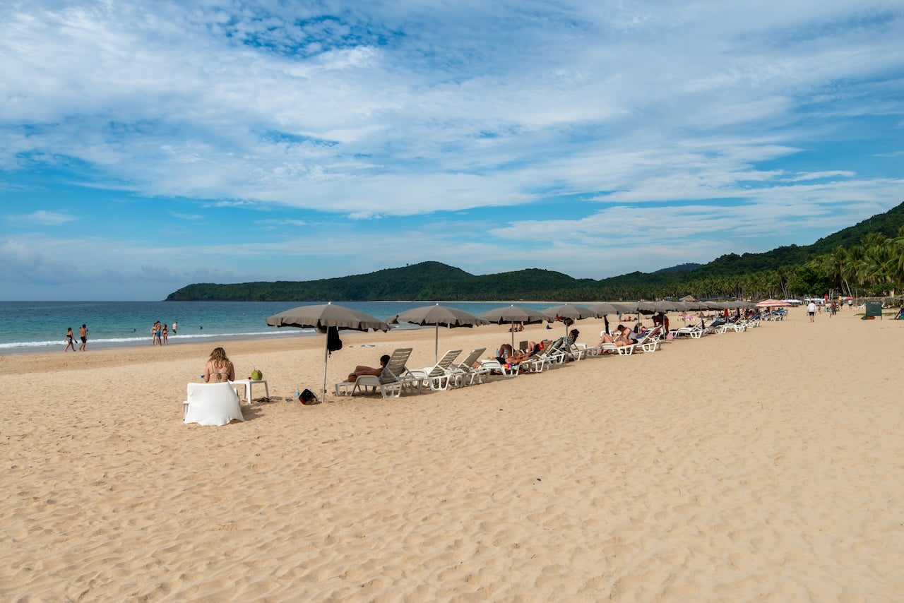 Is Nacpan Beach swimmable? Calm blue water beside a sandy beach with umbrellas and lounge chairs.
